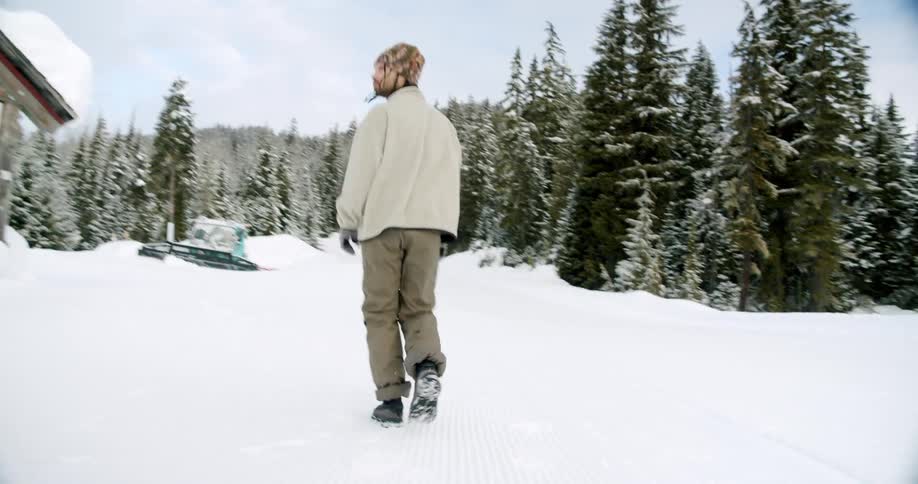 A person in winter clothing walks across a snow covered landscape with a snowy forest in the background