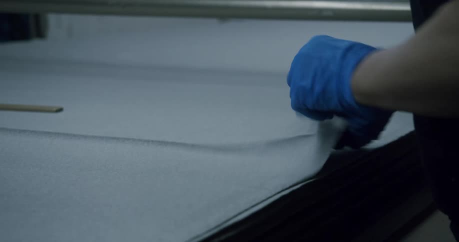 Gloved hands use a wooden ruler to flatten a large piece of cloth over a table