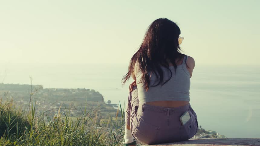 A woman sitting on the floor with a cell phone in her back pocket and she is pointing forward where there is a seascape on the horizon