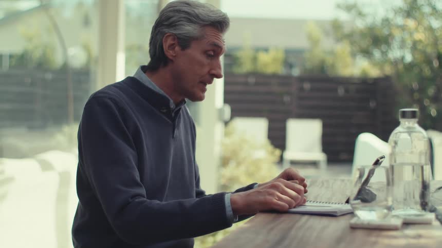 A grayhaired man wearing a blue coat is sitting at an outdoor table looking at a paper and talking to a tablet that is propped up against him