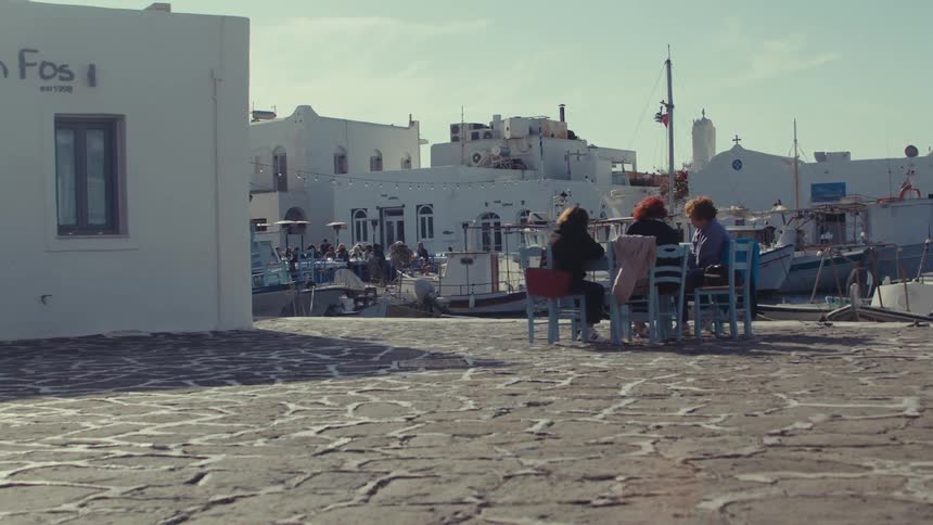 Three women sitting at a table outdoors in a restaurant with other people also sitting on the background next to a pier during daytime