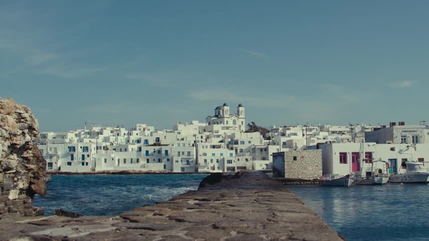 A greek island town with whitewashed buildings and a church dome sits on the coast overlooking a calm harbor