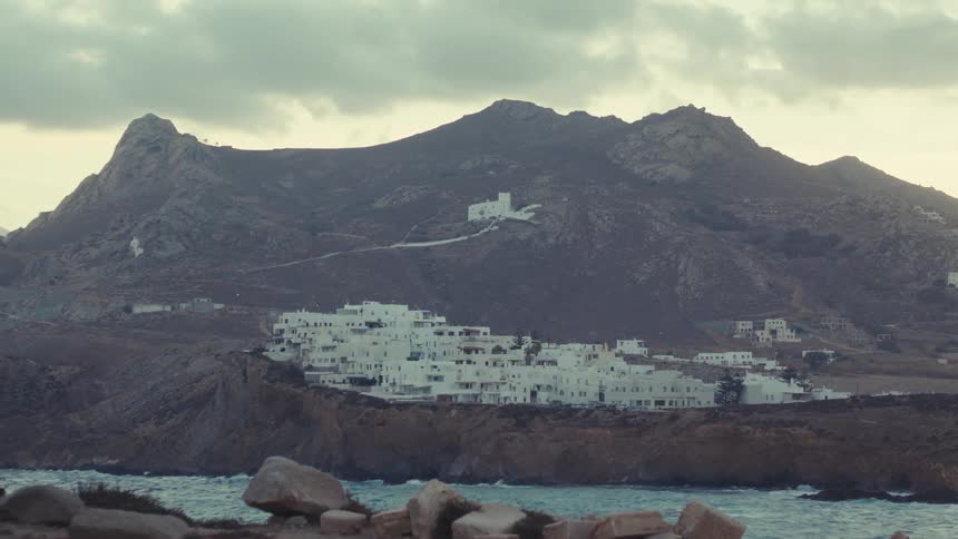 A cluster of whitewashed buildings clings to the side of a rugged mountain under a dramatic cloudy sky
