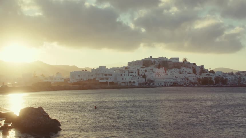 The sun sets over a whitewashed town on a greek island casting a warm glow on the calm waters