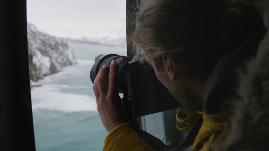 A man in a yellow jacket with a camera pointing outside the room he is in to take a picture