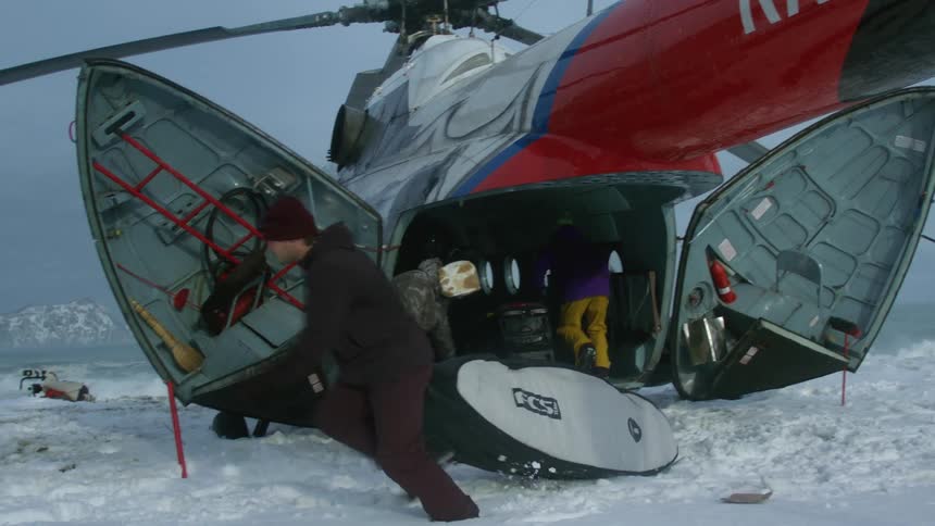 People unpacking a surfboard from a helicopter on a snowy landscape