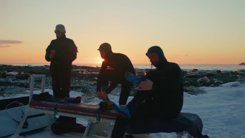 Three individuals are outdoors on a snowy landscape at sunset wearing wetsuits around a sled near the coast