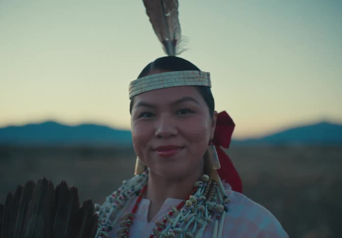 Woman wearing traditional native american attire smiles while standing in a field after dusk
