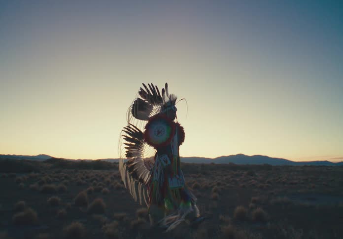Man wearing native american attire walks across scrub land while sunlight brightens the horizon