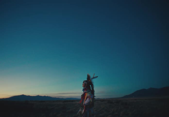 Man wearing native american attire stands in a field after dusk