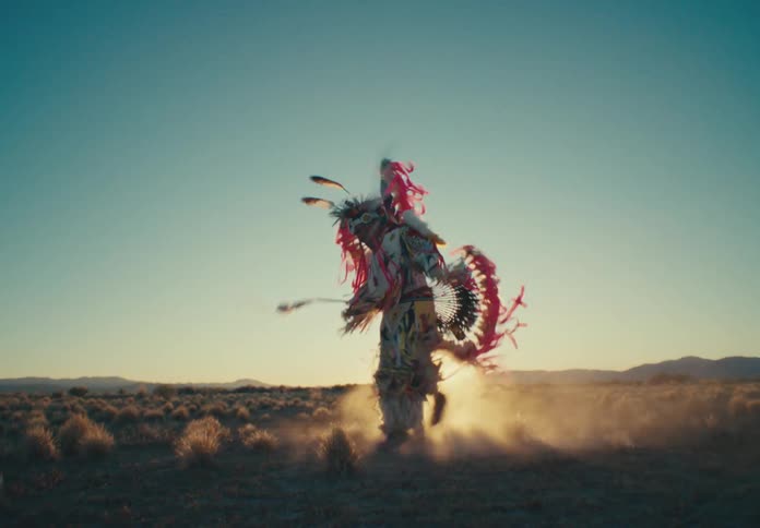 Person wearing a traditional native american dance costume in a desert setting with the sun on the horizon