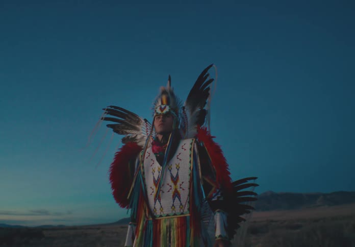 Man wearing native american clothing while standing in a desert environment with sparse vegetation