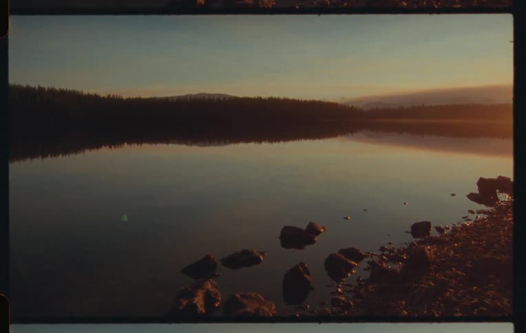 A tranquil lake scene at sunset reflecting the silhouetted trees and sky is shown with rocks in the foreground