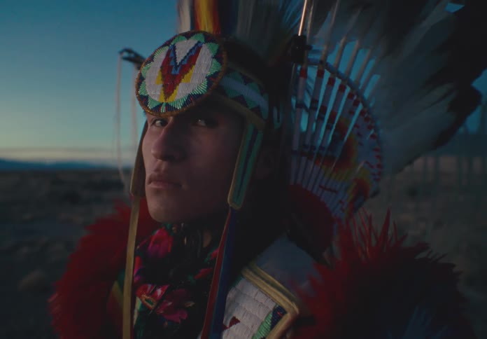 A young native american man in traditional regalia his gaze directed toward the viewer set against a dusky or dawn sky