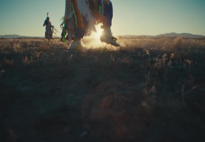 The legs of a person wearing loose clothing full of ribbons who is dancing in a desert region and their feet are kicking up sand