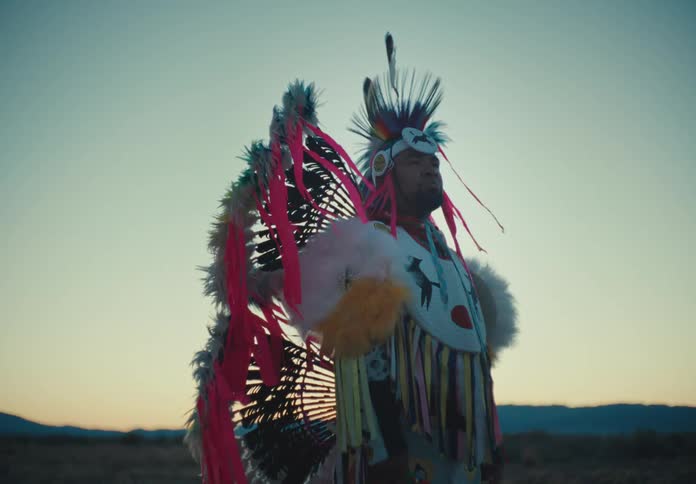 Man wearing traditional native american ceremonial attire stands outdoors in a field on a windy day while red tints the sky above the distant mountains