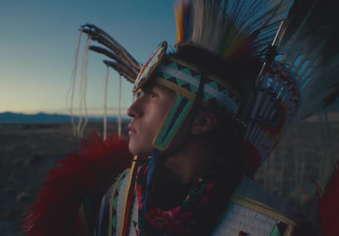 A person is dressed in traditional colorful native american regalia with feathers and intricate patterns standing in an open field