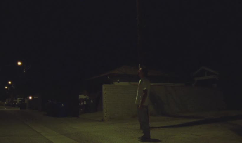 Male youth stands outdoors near a residential road staring up at a streetlight before looking downward