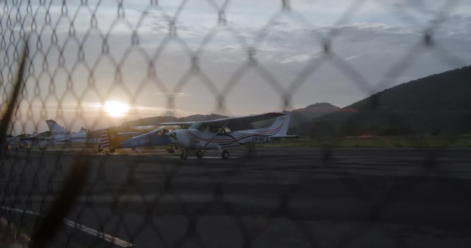 Multiple small airplanes sit at an airfield near the hills on a cloudy day