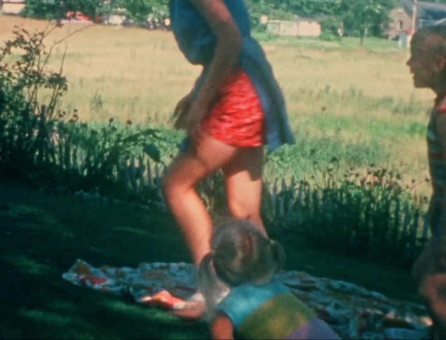 An adult woman is holding the legs of a little blonde girl who is playfully holding her hands in the grass and then letting her go