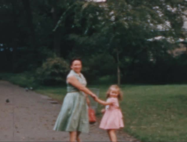 A mother and her little daughter in a pink dress are walking on a concrete path in a backyard with grass and a tree on the right side