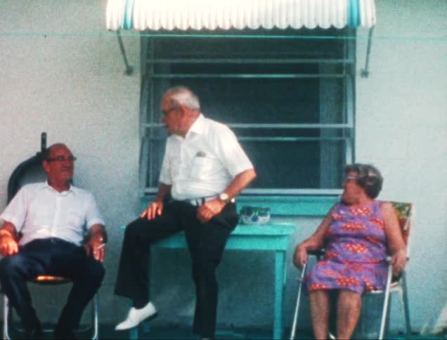 A man and a woman are sitting in blue chairs and then an old man arrives and sits at the table between the two and makes a negative sign with his finger to each of them and then leaves