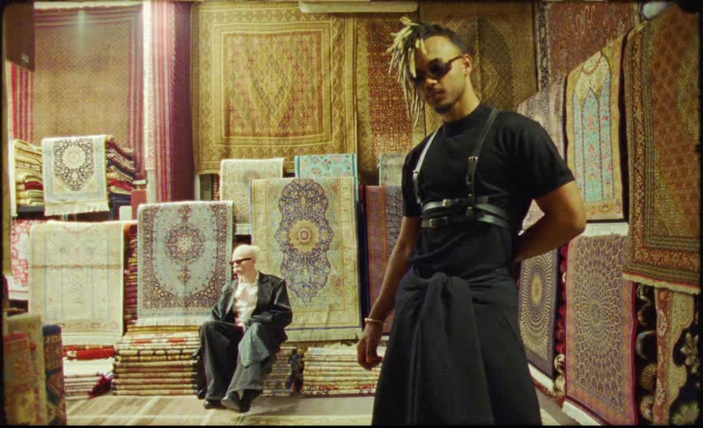 Two men model clothes in front of hanging rugs