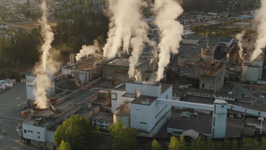 An industrial facility with multiple buildings emits large plumes of white smoke surrounded by trees and a nearby residential area