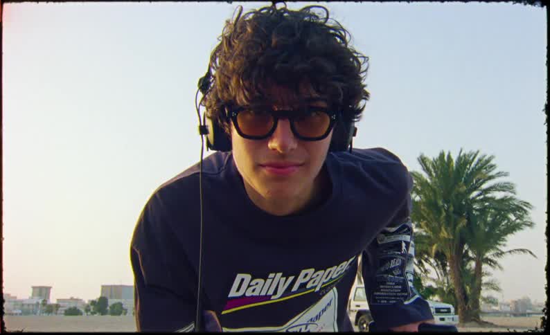 Young man wearing headphones and a graphic longsleeve shirt stands on a dirt lot with a suitcase
