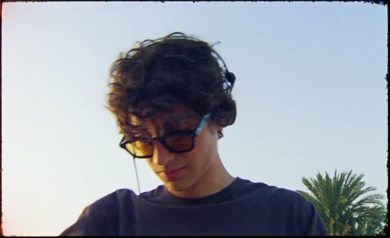 A young man stands in a parking lot with a suitcase and puts on his headphones