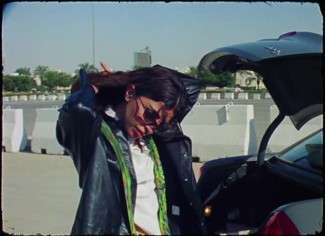 Woman standing next to the open trunk of a car putting on a green tie
