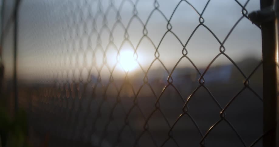 Several small planes are parked on an airfield at sunset seen through a chainlink fence