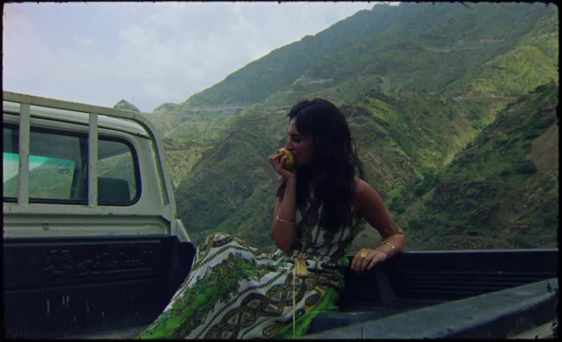 Woman in green dress sitting in bed of a truck eating fruit near a grassy hillside