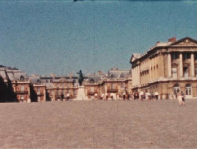 Some people walking in a square with a statue in the center and several buildings behind it
