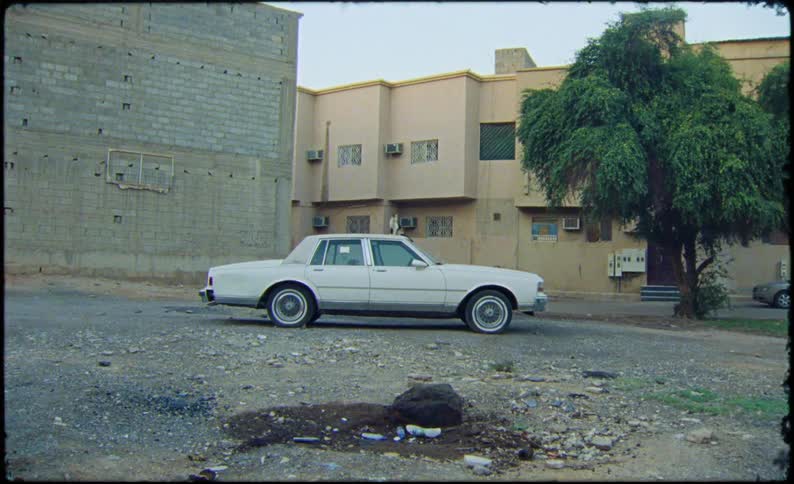 A car passes by in the foreground as the camera zooms out from a closeup shot of a parked white sedan revealing a rugged unpaved lot beige apartment buildings and a large tree in a quiet residential neighborhood