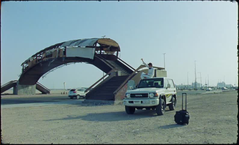 A person is sitting on the hood of a white toyota suv parked on a dirt area next to an arched pedestrian bridge