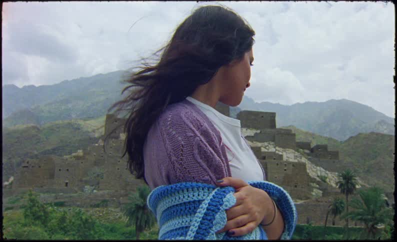 A woman is standing in front of an ancient stone fortress with mountainous terrain holding a blue and white crocheted blanket