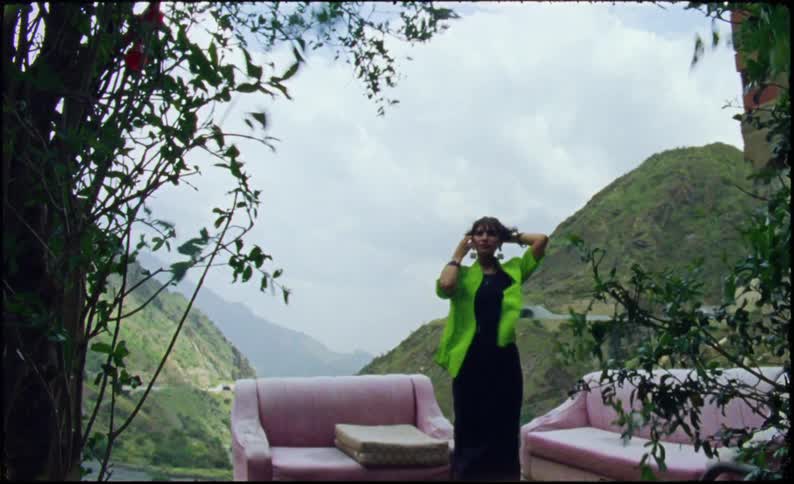 Woman holding her hair while standing on a balcony with pink armchairs and a background of mountains