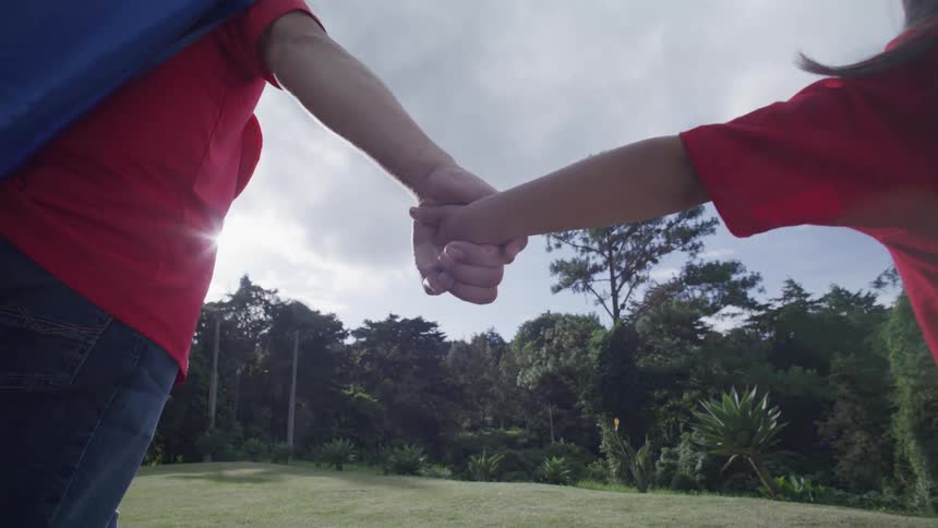 Father and daughter holding hands both wearing a red tshirt and a blue cape in a field