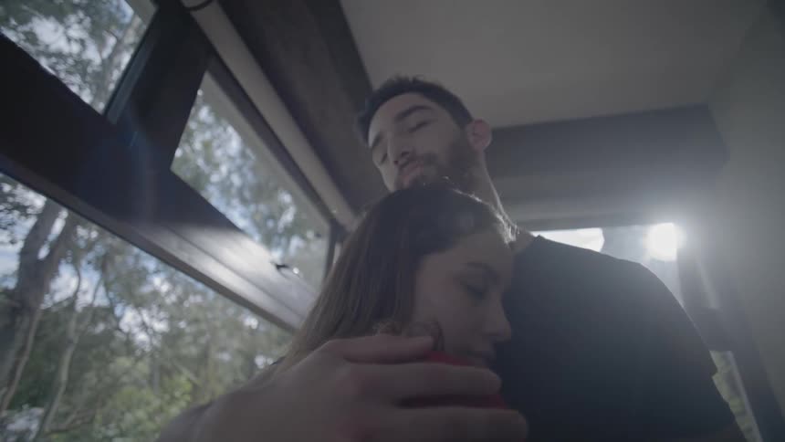A man in a black shirt and a woman in a red shirt embrace with their eyes closed standing next to a window