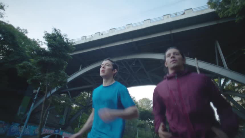 Two men running on the side walk under a bridge