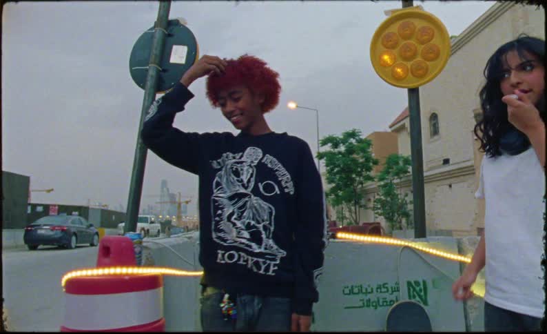 A teenage girl applies chapstick while talking and laughing with her friends near the street