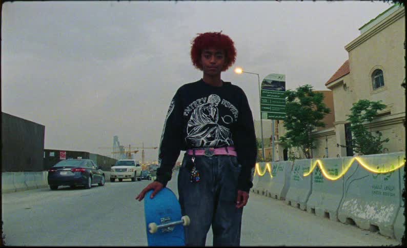 A young woman wearing jeans and a black sweatshirt stands beside the road holding a skateboard ready to ride or take a break from skating
