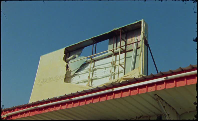 A dilapidated billboard with missing sections leaning against a building with a red roof against a clear blue sky