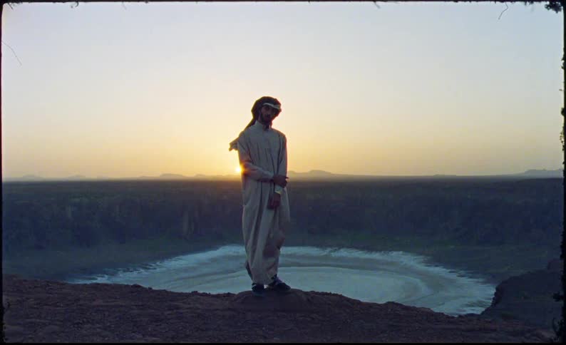 A portrait of a man in loose clothing and a cloth hat standing on the edge of a hill overlooking a natural region with a large crater