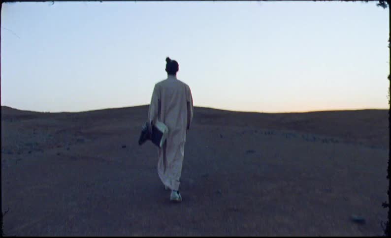 Man wearing a long tunic walks through the desert under a clear sky