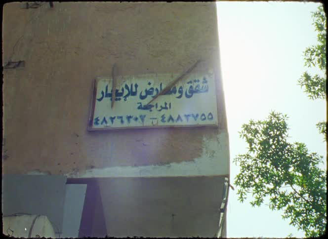 A worn signboard on a wall with arabic text surrounded by sunlight and trees