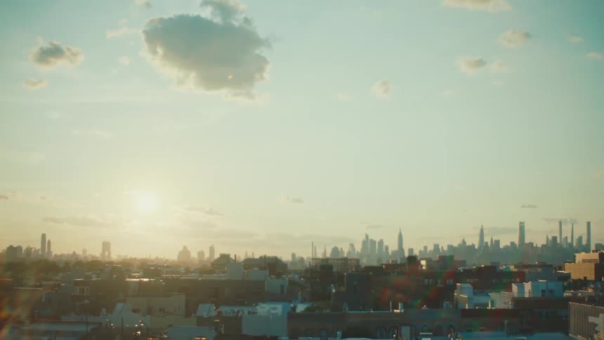 A city skyline at sunset features silhouettes of various buildings and skyscrapers against a warm glowing sky with scattered clouds