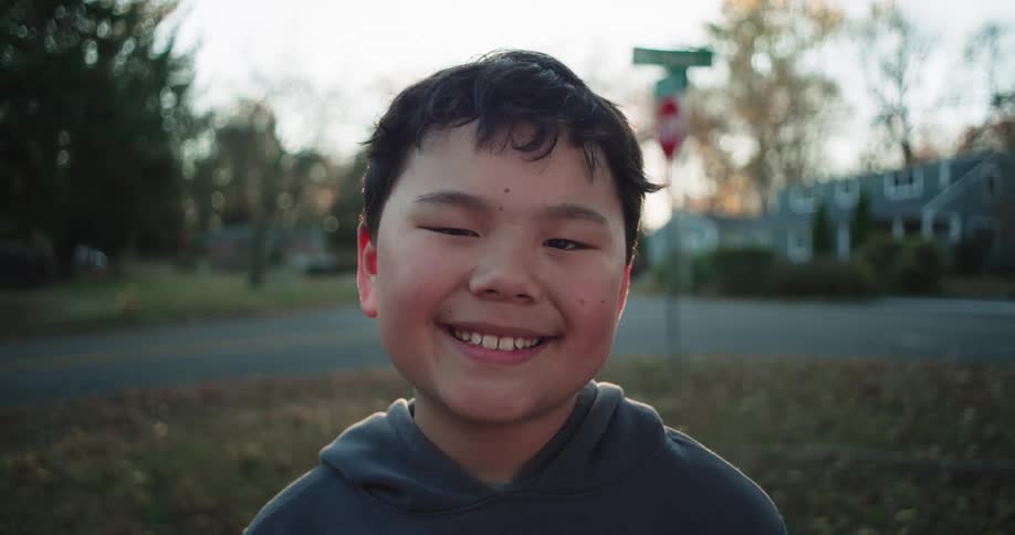 A cheerful boy with a gray hoodie smiling warmly in an outdoor suburban setting during late afternoon with soft sunlight and trees in the background