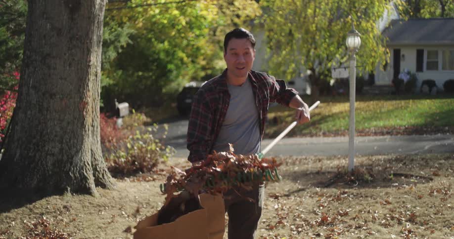 A man is raking autumn leaves into a paper yard waste bag in a residential neighborhood with trees and houses in the background